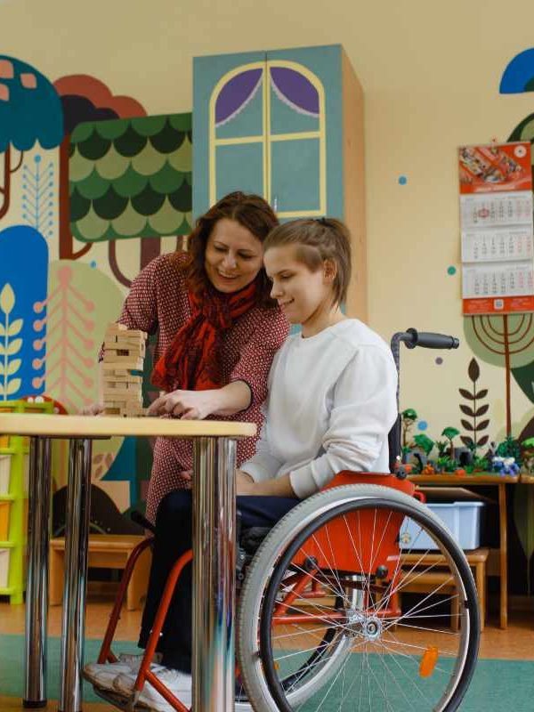 a girl in a wheelchair is playing board games sitting at a table. A female psychologist is engaged with a disabled person.